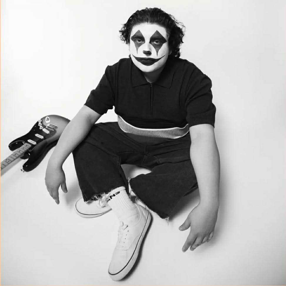 Black and white image of Tony Jupiter, a Chicano surf-goth artist from El Paso, TX. He is wearing clown face paint, wearing black clothes, white shoes and socks and is sitting on the floor next to a guitar to the right of him.