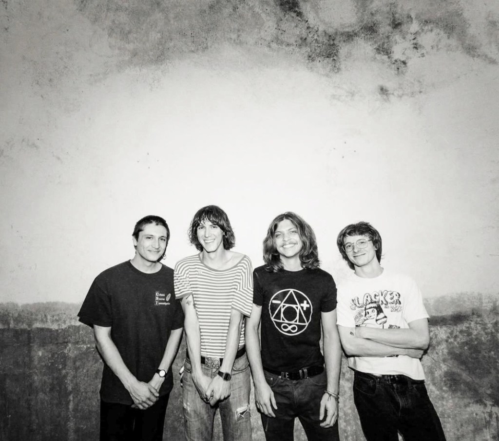 Black and white press photo of Austin-based band Gus Baldwin and the Sketch. The band, comprised of four members dressed in t-shirts and jeans, is standing in front of a nature-inspired background with lots of sky and a cliff.