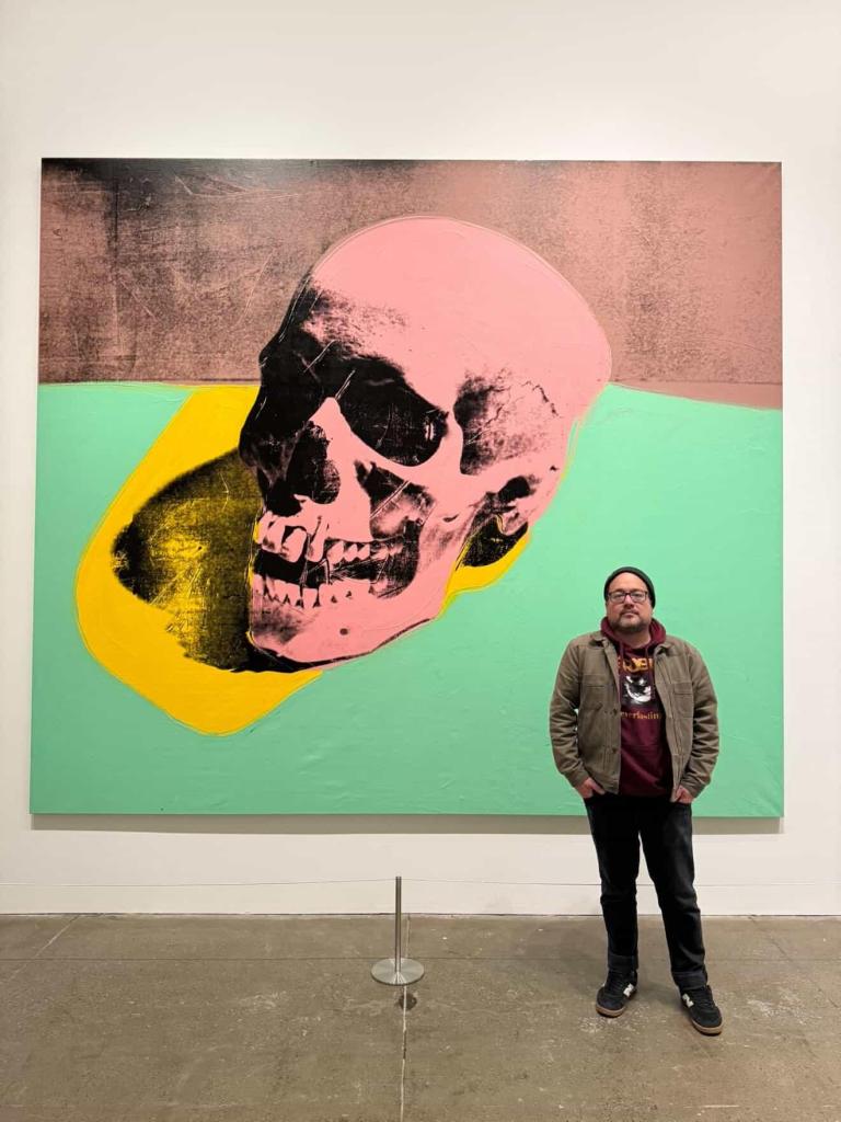 Luis Mota stands in front of one of Andy Warhol's skull prints. The skull is pink with a yellow and teal and pink background.