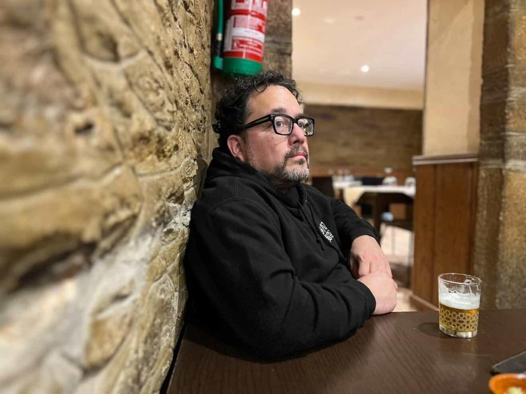 Former El Pasoan Luis Mota sits with his back again a stone wall and he sits at a table with a small glass of beer. He is wearing a black hoodie and black-rimmed glasses and is looking off in the distance.