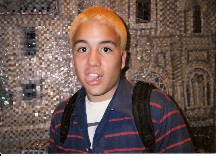 Luis Mota in his backyard punk days in El Paso. He has dyed hair that is orange and is sticking his tongue out as he stares at the camera. He is wearing a navy blue polo with red stripes and a white shirt with gray stripes under it.