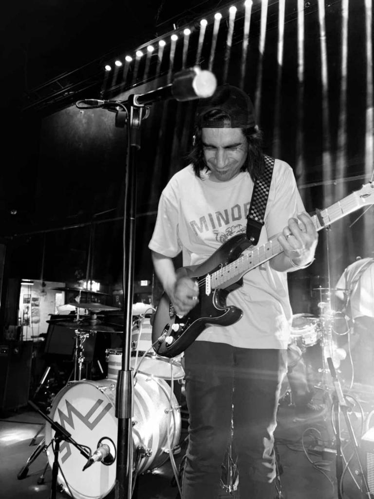 Black and white photo of Adrian Reyes, an El Paso singer/songwriter who goes by the name My Darling May, as he plays guitar. He is in a studio and standing in front of a drum set and in front of a microphone stand.