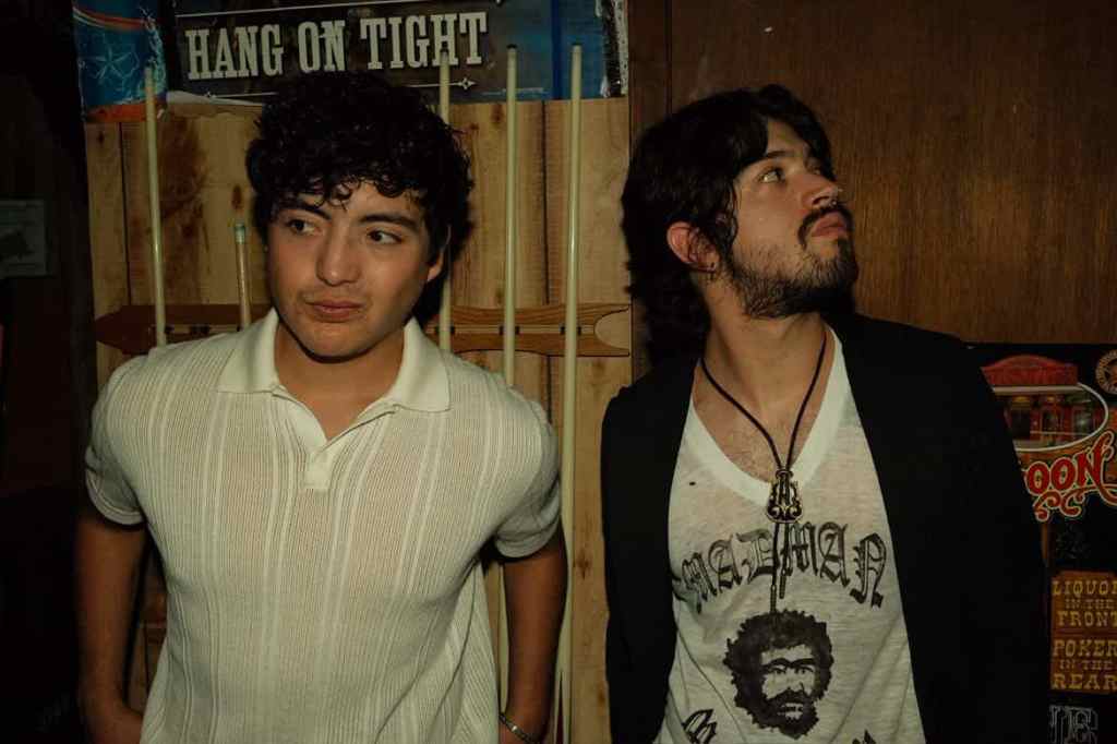 Band photo of The Veronas, a rock and roll band from El Paso, Texas. The picture is of two of the bands members standin in front of a stand for pool sticks in a bar. One member is wearing a white shirt and is looking to his righ and the other band member is wearing a white t-shirt and black sweater and is looking off to their left.