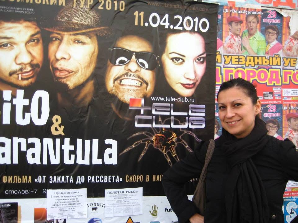Photo of Olivia "Tina" Torres in all black attire standing in front of a poster announcing a show of Tito & Tarantula, whom she is a tour manager.