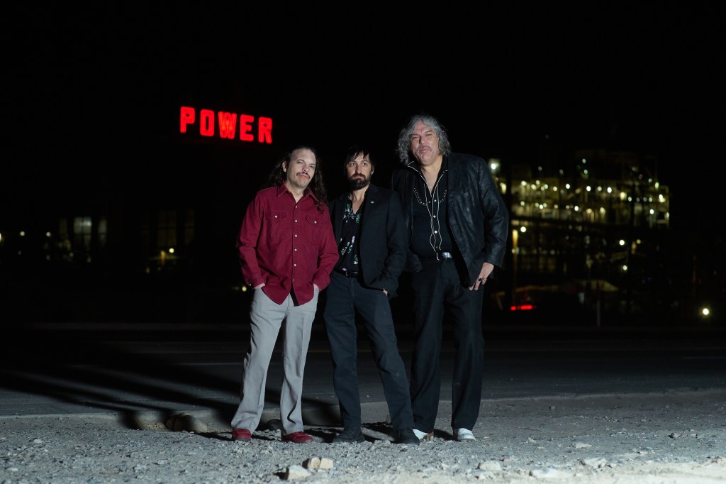 Photo of El Paso, TX, band Krunchies, a garage rock power trio comprised of Wally Byers, Ernie Carrillo, and Carlos Palacios, standing in a dirt lot with the El Paso Electric - Rio Grande Power Plant red "Power" sign in West El Paso in the background.